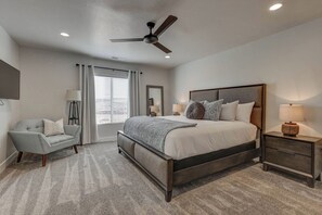 Une chambre à coucher comprenant un lit, des tables de chevet, une chaise, un ventilateur de plafond et une fenêtre avec des stores.