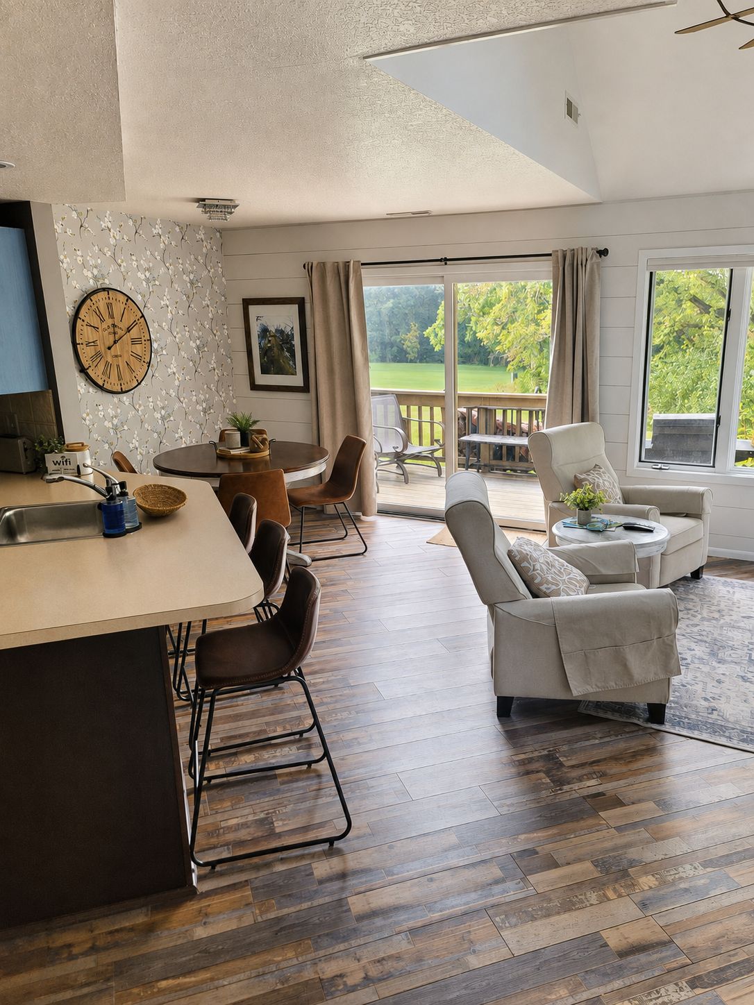 A modern kitchen with a wooden island, dining chairs, a sofa, and a view of a garden through large windows.