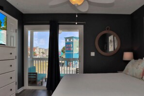 A bedroom with a bed, a dresser, a mirror, and a view of a beach and buildings. - Mystic Nights By The Sea (Port St. Joe)