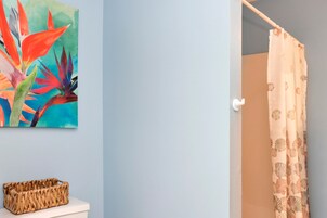 A bathroom with a shower curtain, a toilet, a woven basket, and a colorful painting on the wall.
