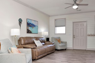 A living room with a brown leather sofa, a grey armchair, a painting, a ceiling fan, and a door.
