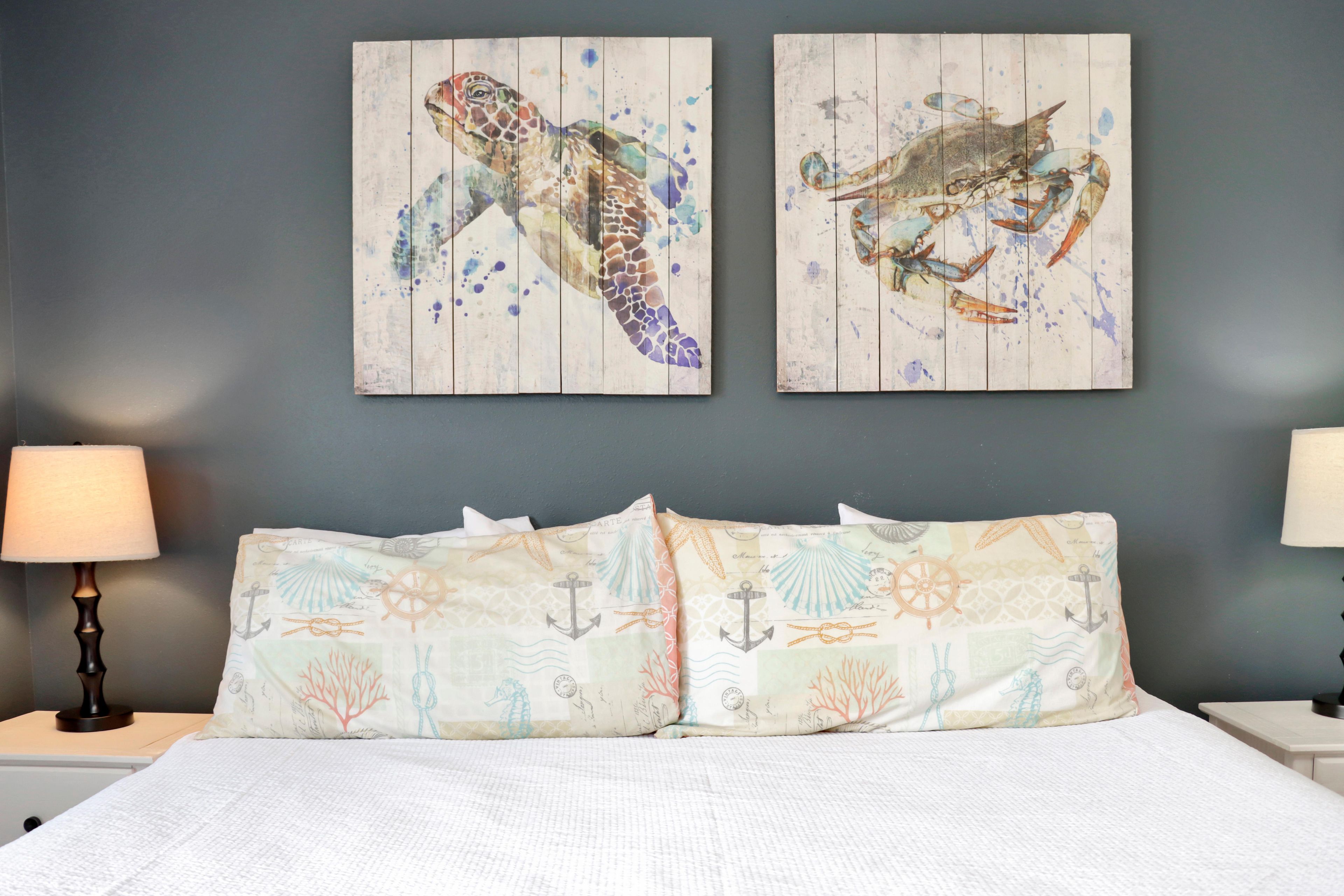 A bedroom with a bed, two wall-mounted paintings of sea creatures, and a nightstand with a lamp.