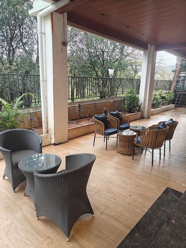 An outdoor patio area with wicker furniture, a glass table, and a view of a garden with a metal fence.