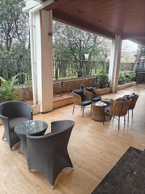 An outdoor patio area with wicker furniture, a glass table, and a view of a garden with a metal fence.