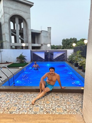 Ein Mann sitzt an einem Schwimmbecken mit Fliesenboden, einer Poolleiter und einem Gebäude mit Rundbogenfenstern im Hintergrund.