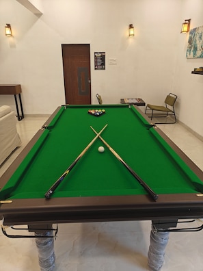 A pool table with a cue ball and two cues, a chair, and a chessboard in a room with a door and wall-mounted lights.
