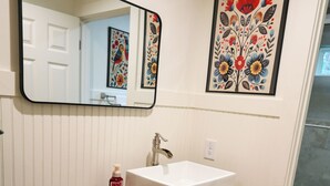 A bathroom with a wooden vanity, a white sink, a mirror, and a framed floral artwork on the wall. - The Loft at Goshen Falls (Clarkesville)