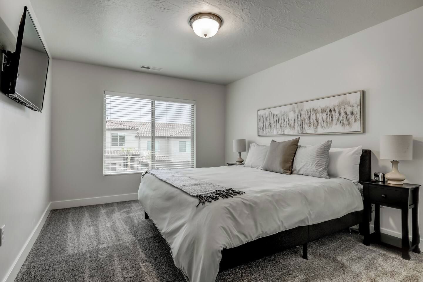 A bedroom with a bed, a nightstand, a lamp, a wall-mounted TV, and a window with blinds.