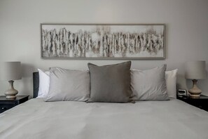 Un lit avec un oreiller gris et des oreillers à rayures, un mur uni orné d’un tableau, et deux tables de chevet avec des lampes.