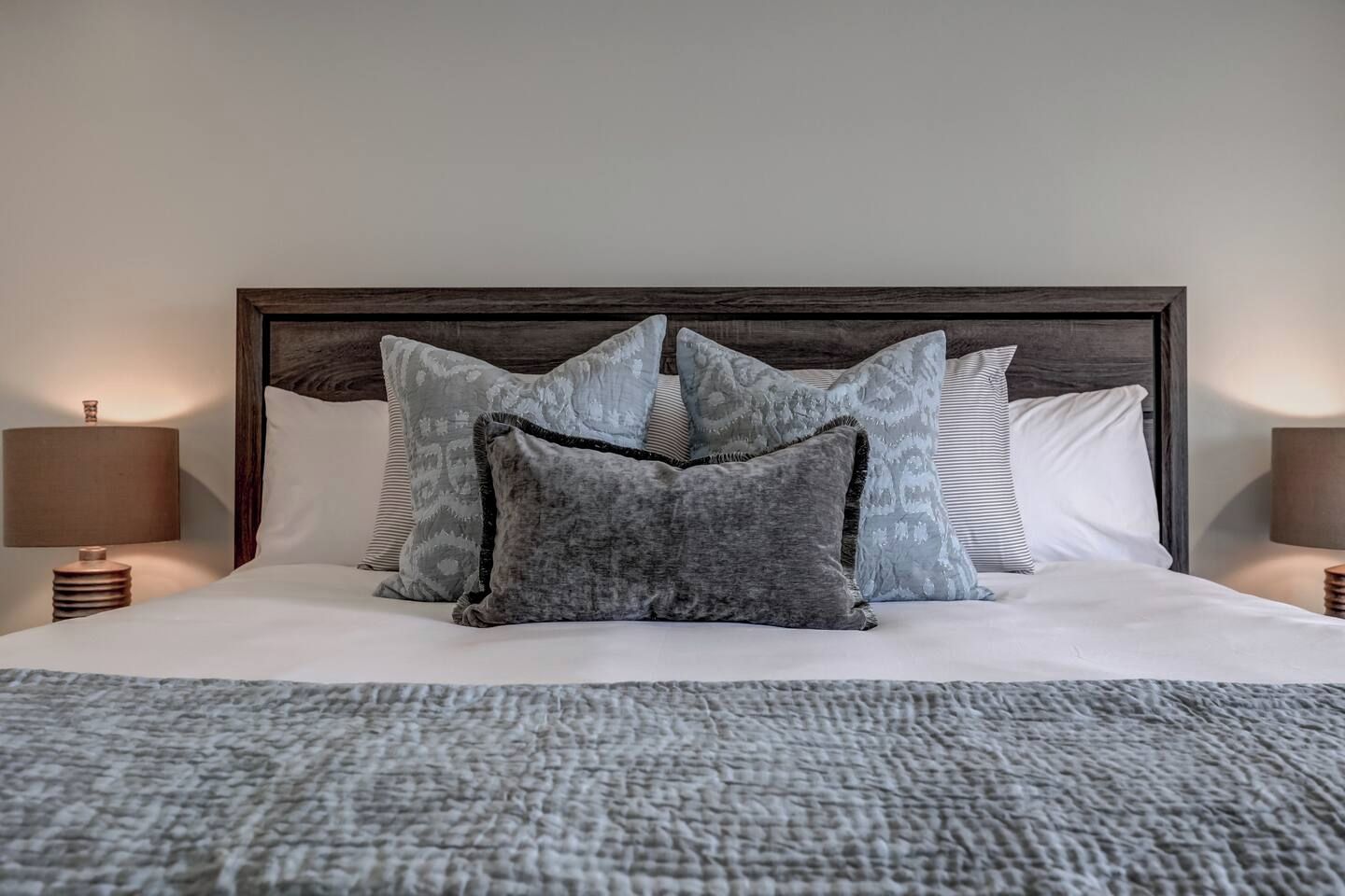 A neatly made bed with a grey textured blanket, a dark wooden headboard, and multiple pillows in shades of grey and white.
