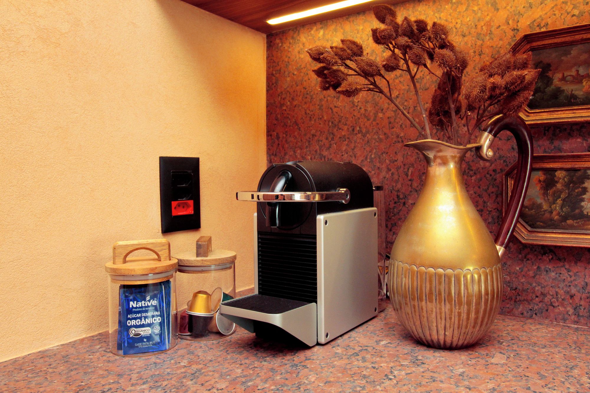 A coffee machine, a container of organic sugar, a metal pitcher, and a small wooden stool on a kitchen counter.