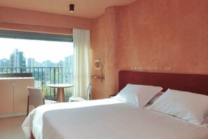 A hotel room with a bed, a small table, and a chair. There is a window with a view of the city. - MOBE Studios (São Paulo)