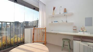 Um apartamento moderno com uma janela ampla, uma cadeira de madeira, um banquinho pequeno, uma escrivaninha branca e um radiador.