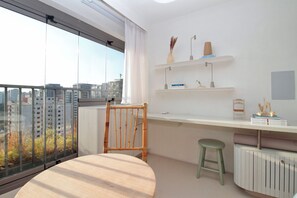 Un appartamento moderno con una grande finestra, una sedia di legno, uno sgabello, una scrivania bianca e un termosifone.