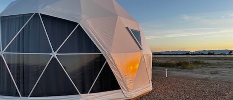 A geodesic dome tent with a clear window, set on a gravel surface with a fence and distant mountains in the background.