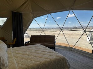 A tent with a bed, a chair, and a window offering a view of a dry landscape.