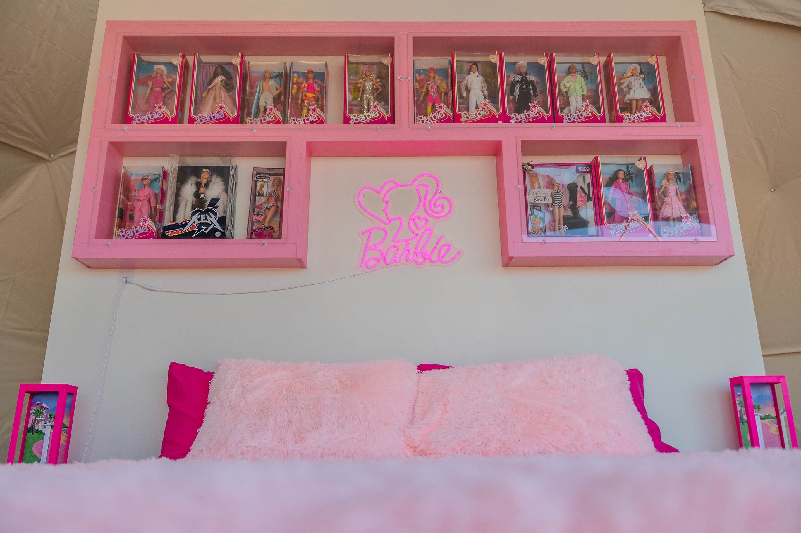 A bedroom with a pink bed, multiple framed pictures on the wall, and a neon sign.