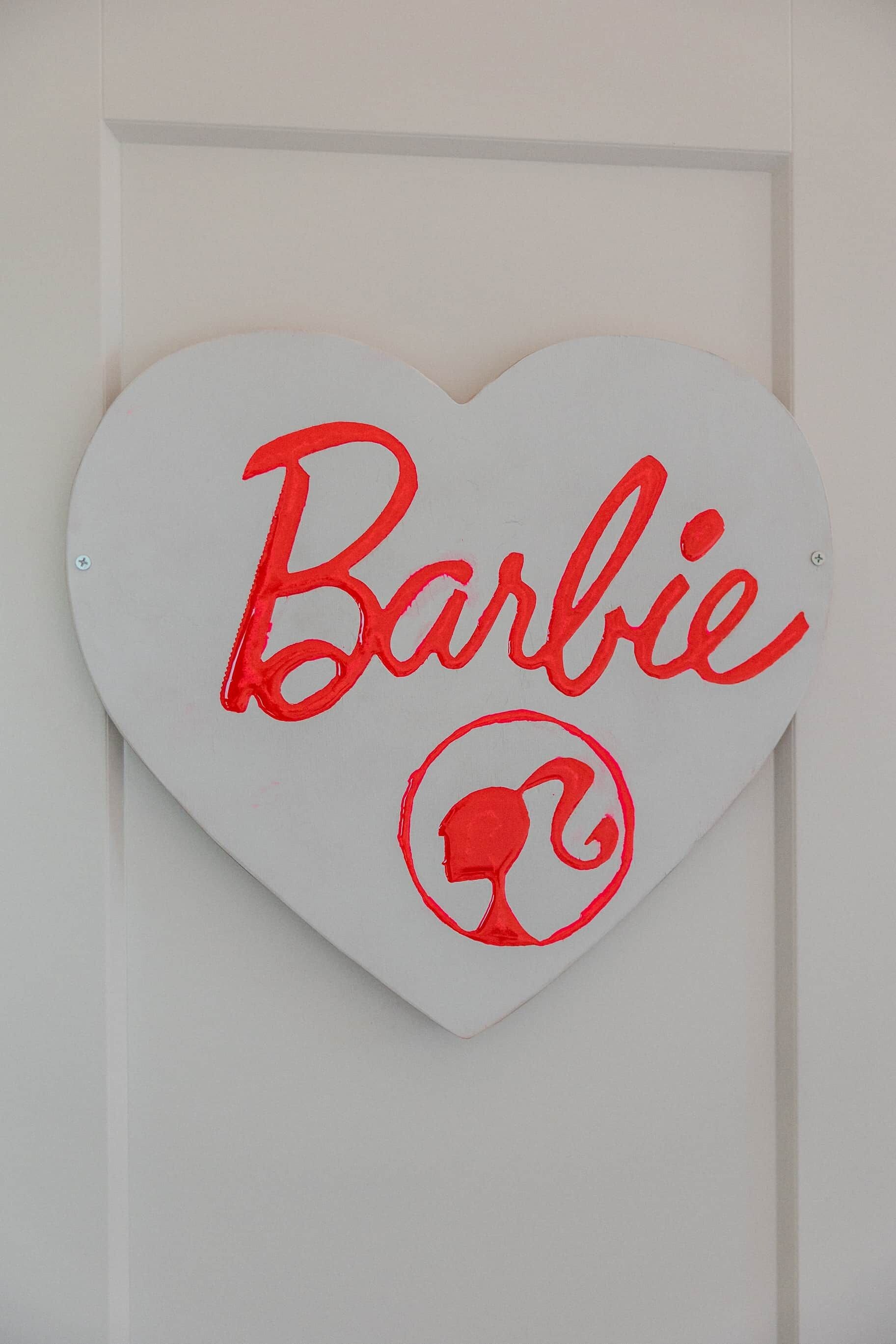 Un panneau blanc en forme de cœur, portant le mot « Barbie » et un logo, fixé sur une porte.