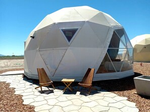 Una estructura de domo geodésico con un ventanal triangular, dos sillas de madera y una pequeña mesa al frente.