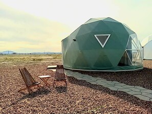 A geodesic dome structure with a triangular window, surrounded by a gravel area with two wooden chairs and a small table.