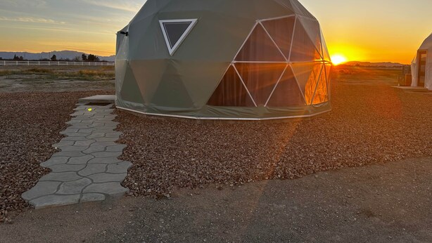 A geodesic dome structure with a triangular window design, situated on a gravel surface with a paved pathway leading to it. - The Beer Dome at Tirrito Farm (Cochise County)