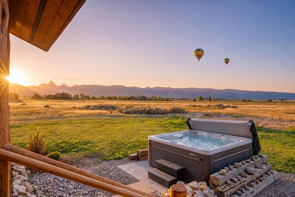 A hot tub with a view of a field and mountains, two hot air balloons in the distance, and a wooden structure in the foreground.