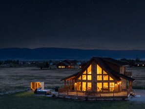 A lodge with a large deck and multiple windows, surrounded by a grassy field and mountains in the background.