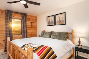 Ein schlichtes Schlafzimmer im Chalet-Stil mit Bett, einem Fenster mit Vorhängen, einem Deckenventilator, einem Nachttisch mit Lampe und zwei gerahmten Bildern an der Wand.