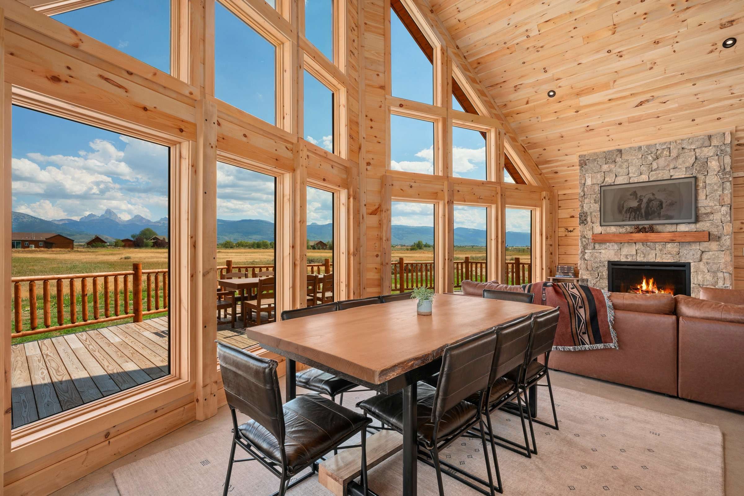 A spacious room with a large wooden table and chairs, a stone fireplace, and expansive windows offering a view of the outdoors.