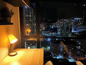 A night view from a window overlooking a cityscape with illuminated buildings, a lamp on a table, and a potted plant on the sill. - Tranquil 1-bedroom apartment in vibrant Bogotá D.C (Bogotá, D.C.)