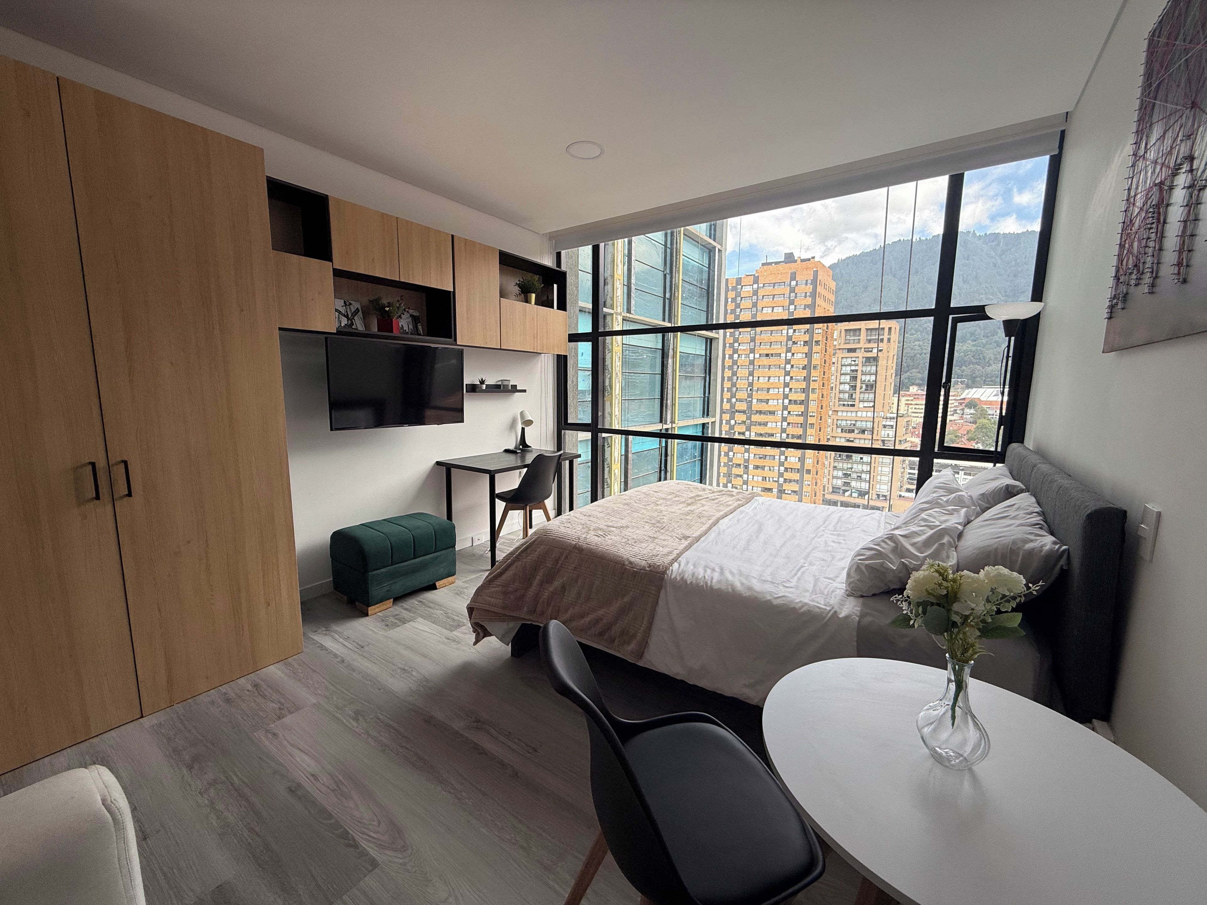 A modern bedroom with a bed, a desk, a chair, and a view of a cityscape through large windows.