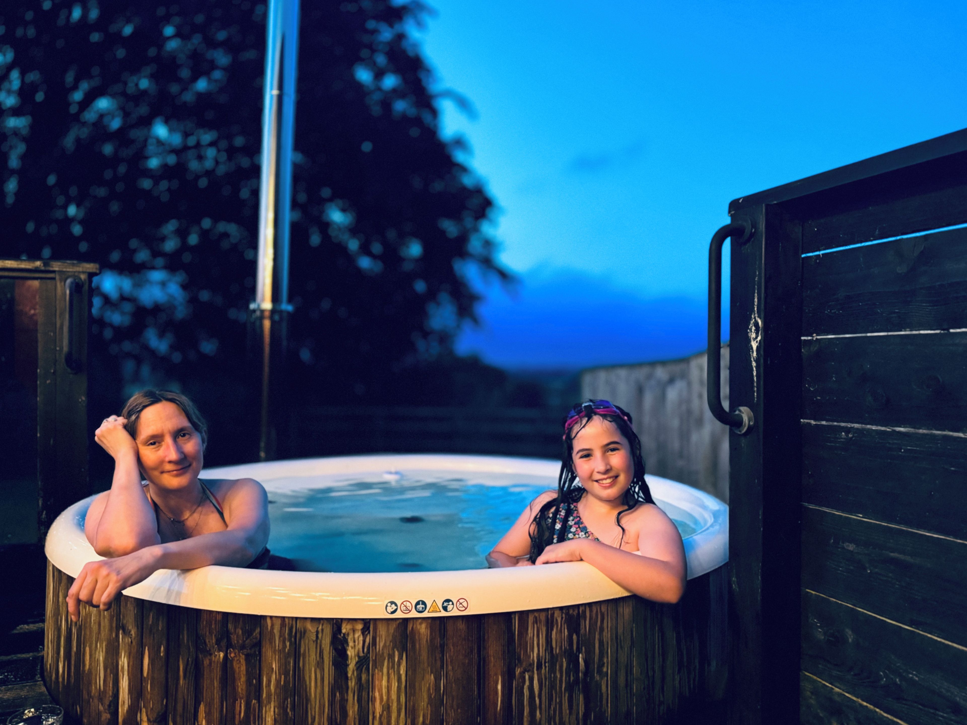 Two people in a hot tub outdoors with a wooden structure and a tree in the background.