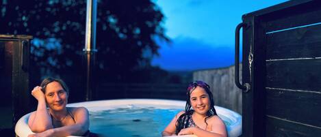 Two people in a hot tub outdoors with a wooden structure and a tree in the background.