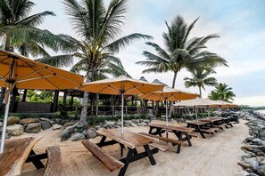 En restaurant ved stranden med udendørs siddepladser, palmer og en klippefyldt kystlinje.