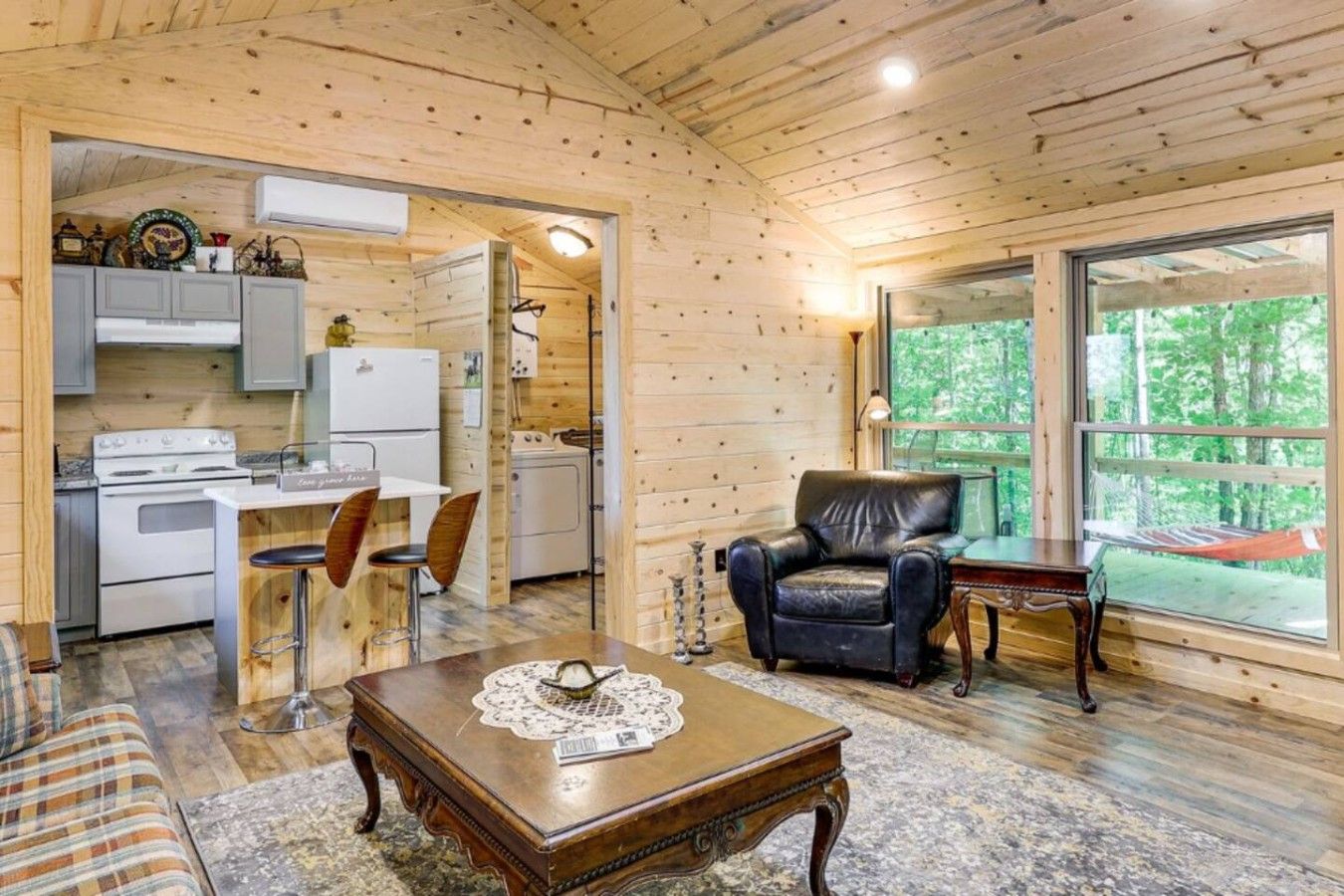 A wooden cabin interior with a kitchen, dining area, and a large window offering a view of the outdoors.