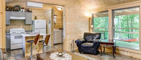 A wooden cabin interior with a kitchen, dining area, and a large window offering a view of the outdoors.