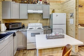 A compact kitchen with a stove, microwave, and refrigerator, wooden walls, and a table with a decorative sign. - Boudreaux Run Cabin Ouachita National Forrest (Langley)
