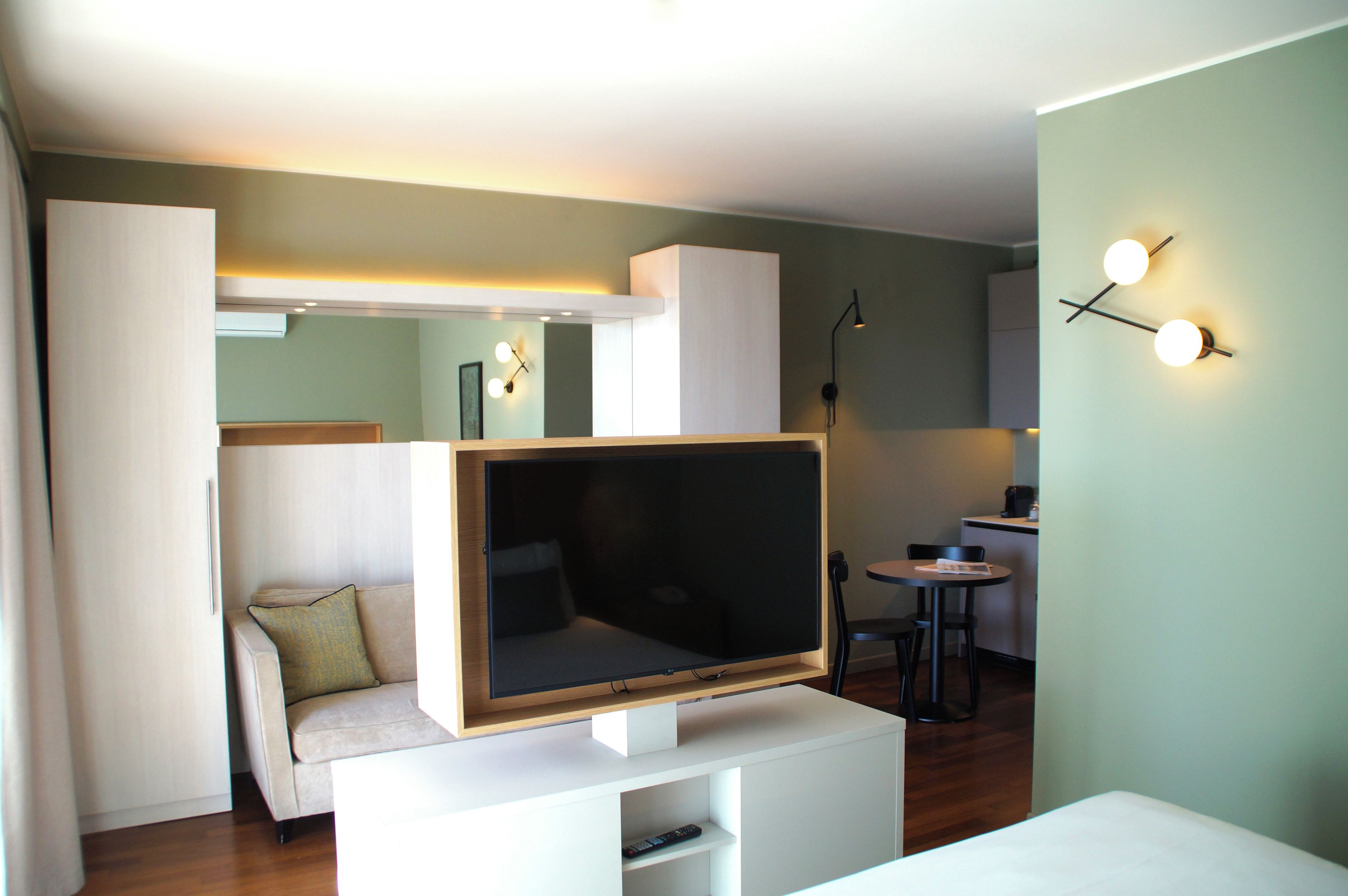Una sala de estar moderna con un televisor de pantalla plana grande sobre un soporte blanco, un sofá con un cojín, una mesita con una silla y un accesorio de luz fijado a la pared.