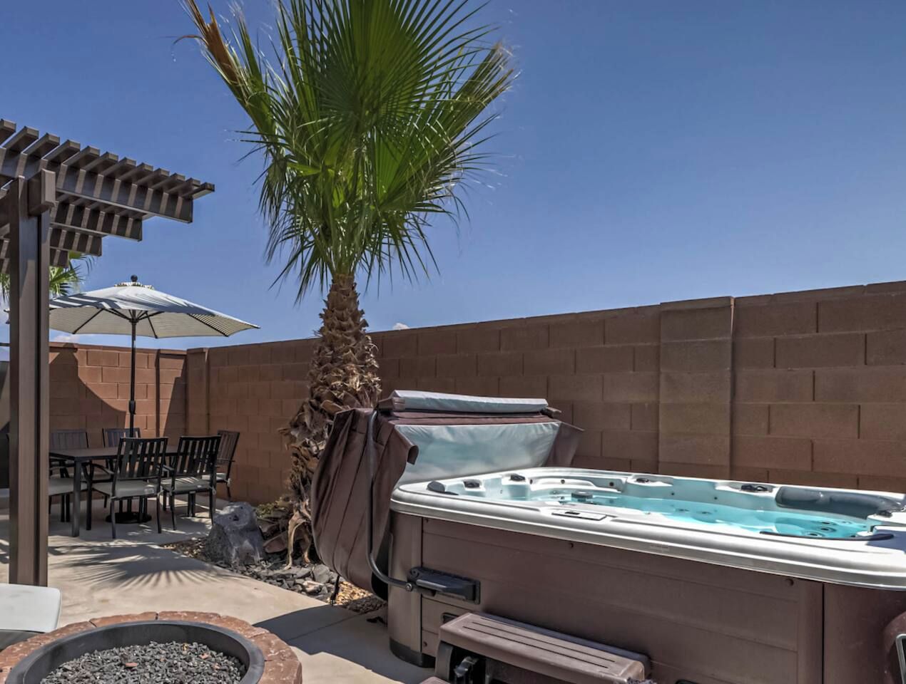 Un jacuzzi en un patio trasero con una mesa y sillas, una palmera y un muro de ladrillo.