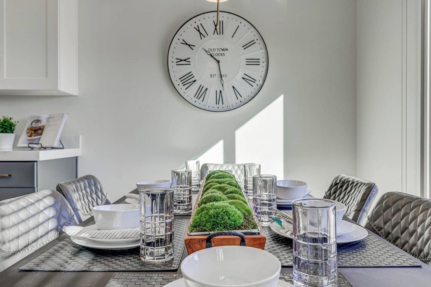 Uma sala de jantar com relógio na parede, mesa posta com pratos, copos e uma planta decorativa.