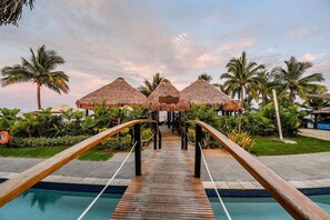 A wooden pathway leading to thatched-roof structures, surrounded by palm trees and greenery. - Denarau | Group Island Paradise | Full Kitchen & Balcony | 2BR (Nadi)