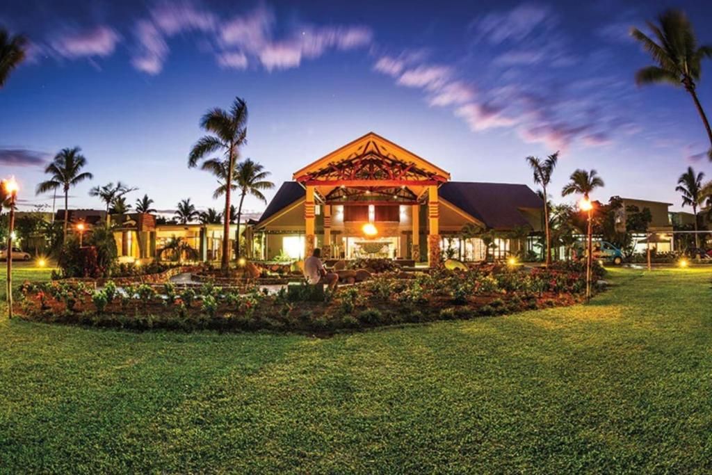 A hotel with a central building, surrounded by a well-maintained lawn and palm trees.