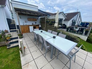 A patio with a long table and chairs, a small kitchen area, and a staircase leading to an upper level. - Helston 5-bed House, Sleeps 12 With Garden (Helston)
