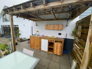 A wooden outdoor kitchen area with a built-in refrigerator, a sink, and a wooden cabinet above. - Helston 5-bed House, Sleeps 12 With Garden (Helston)