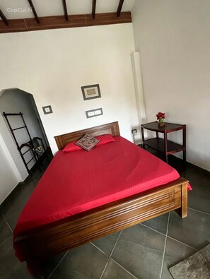 Ein Schlafzimmer mit einem hölzernen Bett, einer roten Tagesdecke, einem kleinen Nachttisch mit einer Vase und einer an der Wand stehenden Leiter.