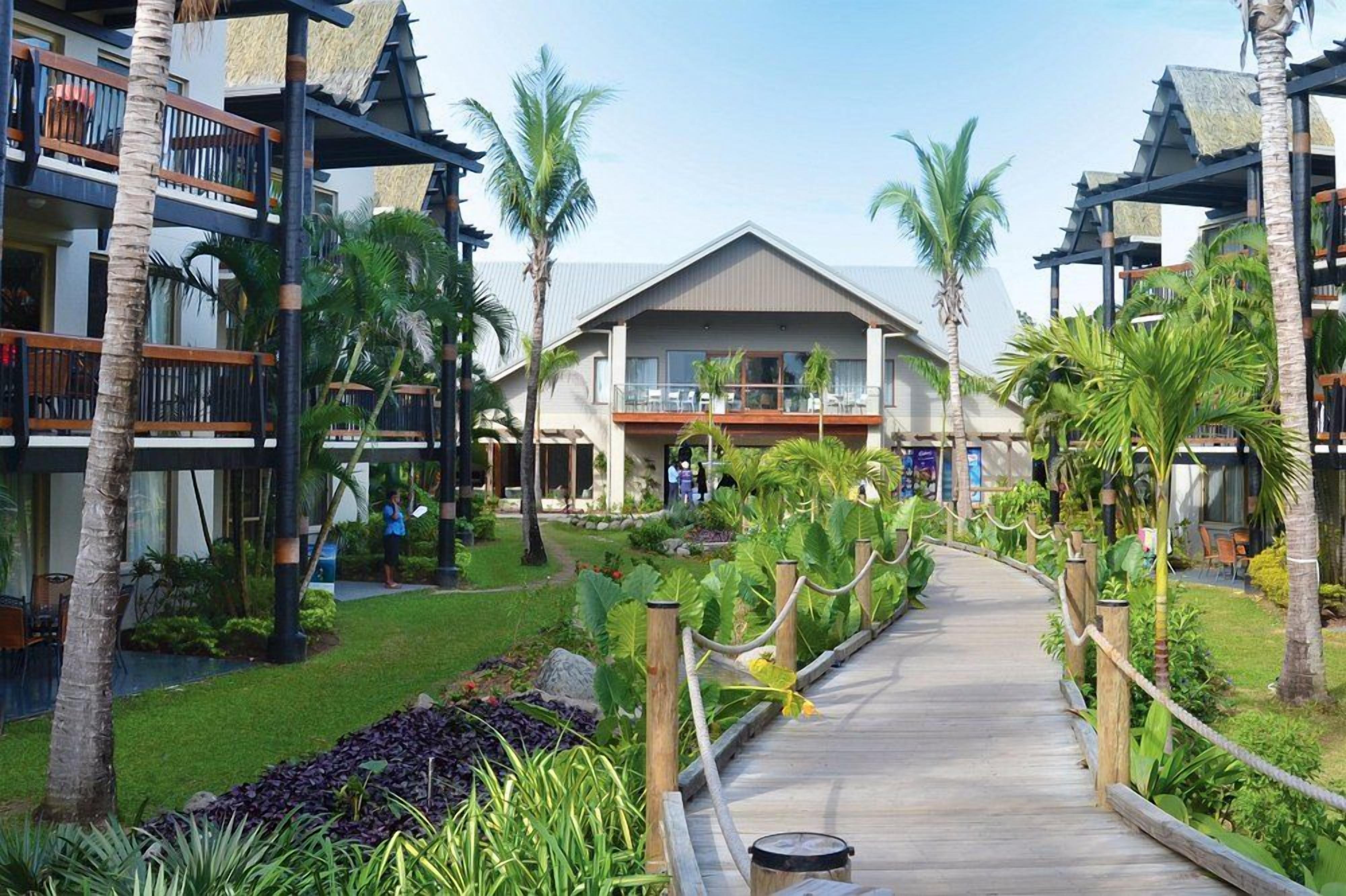 A resort with a central building, multiple balconies, and a wooden pathway surrounded by tropical vegetation.