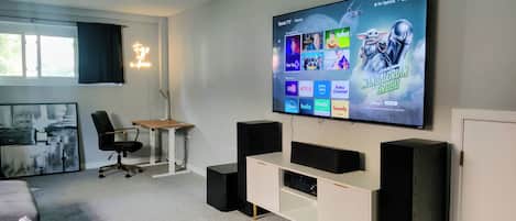 A modern living room with a large flat-screen TV displaying various streaming services, a comfortable seating area with a desk and chair, and a cozy couch.
