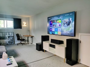 Een moderne woonkamer met een grote flatscreen-tv waarop diverse streamingdiensten worden weergegeven, een comfortabele zithoek met een bureau en stoel, en een gezellige bank.