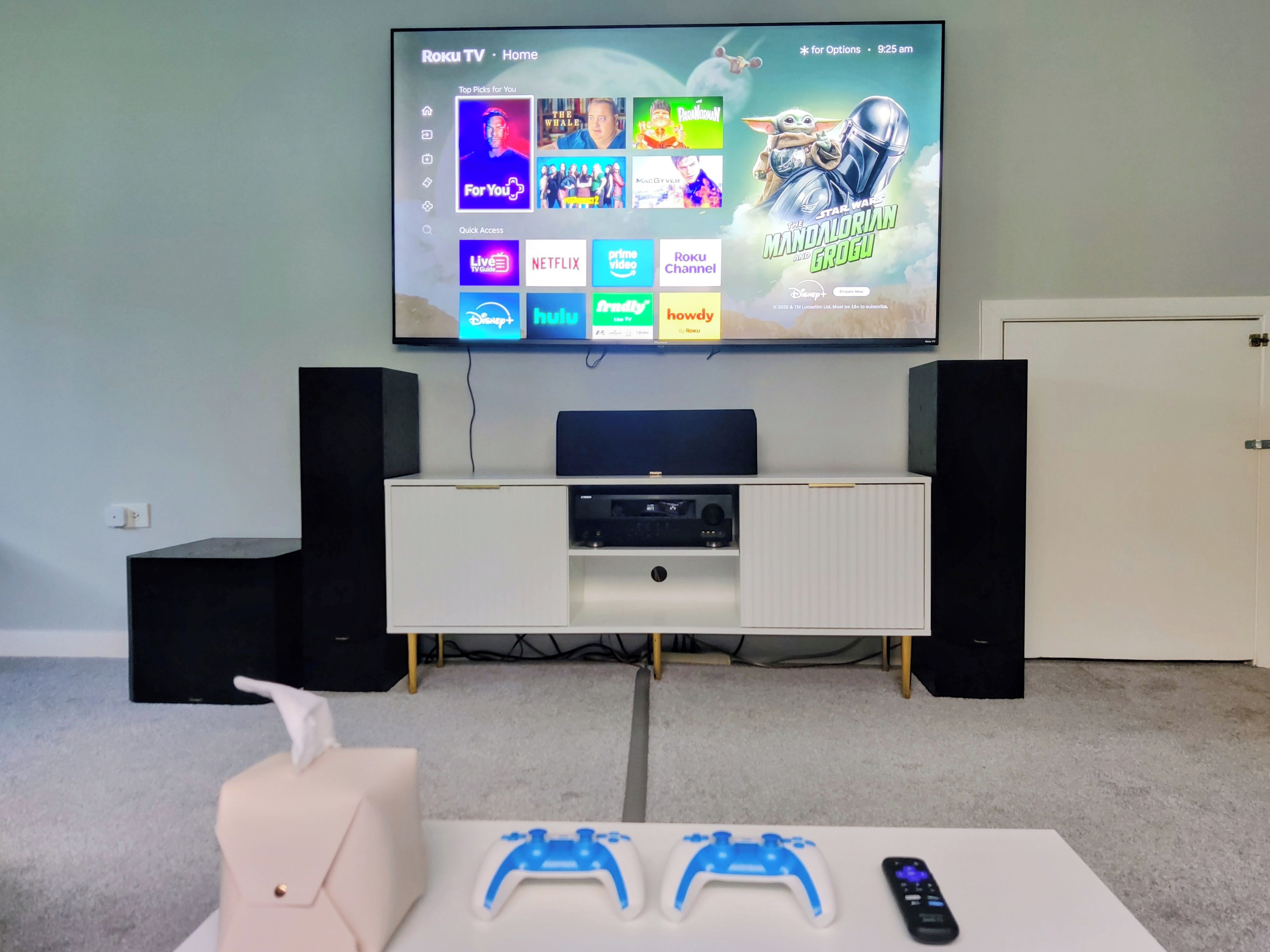 A living room with a flat-screen TV mounted on the wall, a white entertainment unit below, two speakers, and a gaming setup with two controllers and a remote.