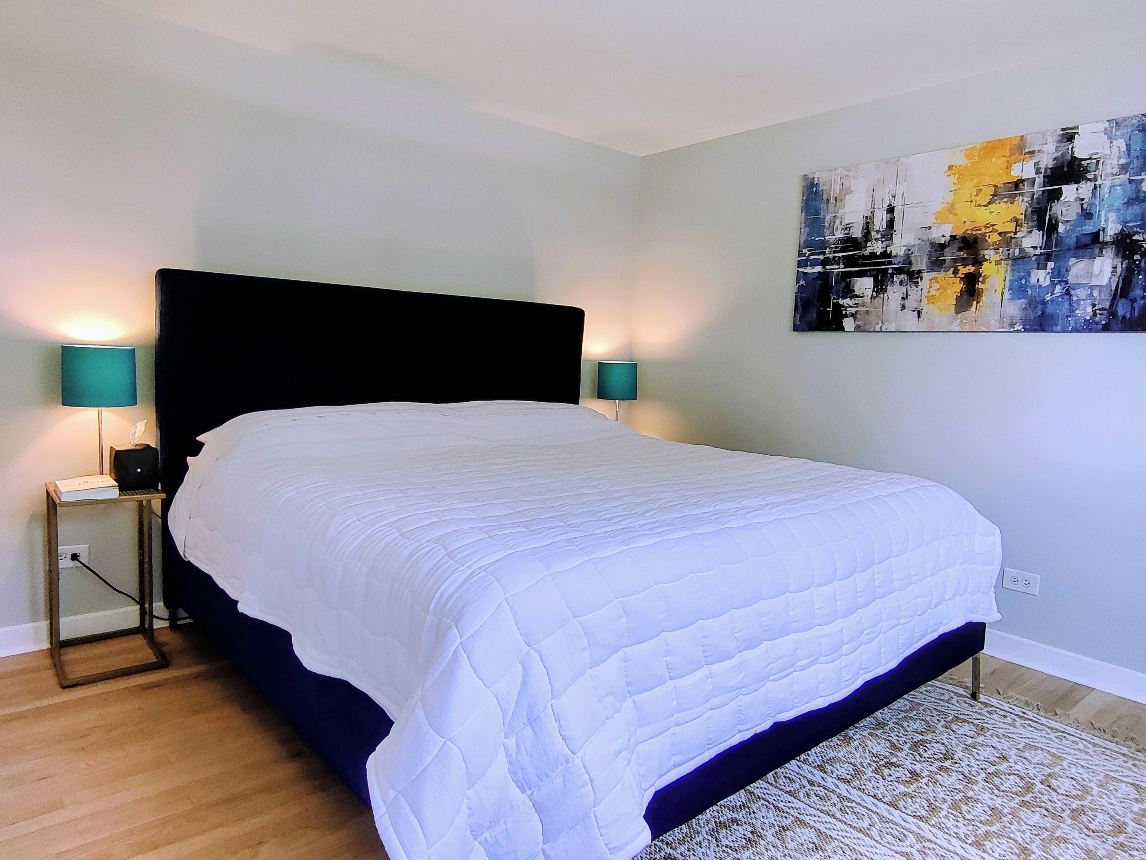 A bedroom with a large bed, two bedside lamps, a nightstand, a wall painting, and a patterned rug.